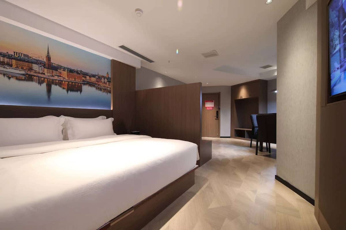 A modern hotel room with a large bed, a cityscape painting on the wall, and a television screen.