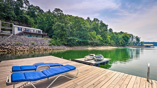 A wooden deck with blue lounge chairs, a dock with boats, and a house with a balcony overlooking a lake.