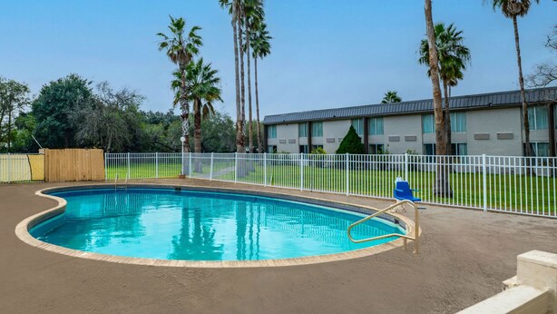 A swimming pool with a curved edge, surrounded by a white fence, palm trees, and a building with a flat roof in the background. - Quality Inn Alice (Alice)