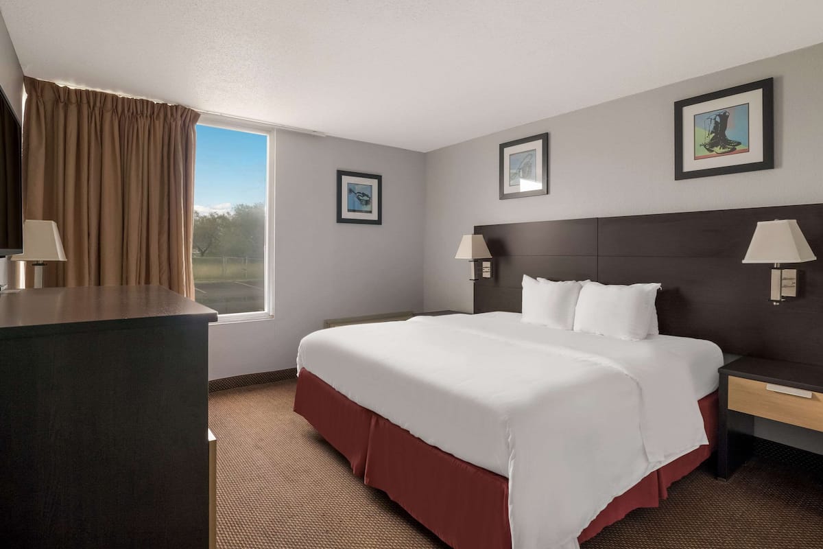 A hotel room with a large bed, two bedside tables with lamps, a window with curtains, and two framed pictures on the wall.