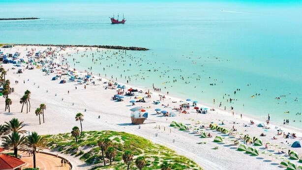 A beach with numerous umbrellas and people, a lifeguard tower, and a ship in the distance.