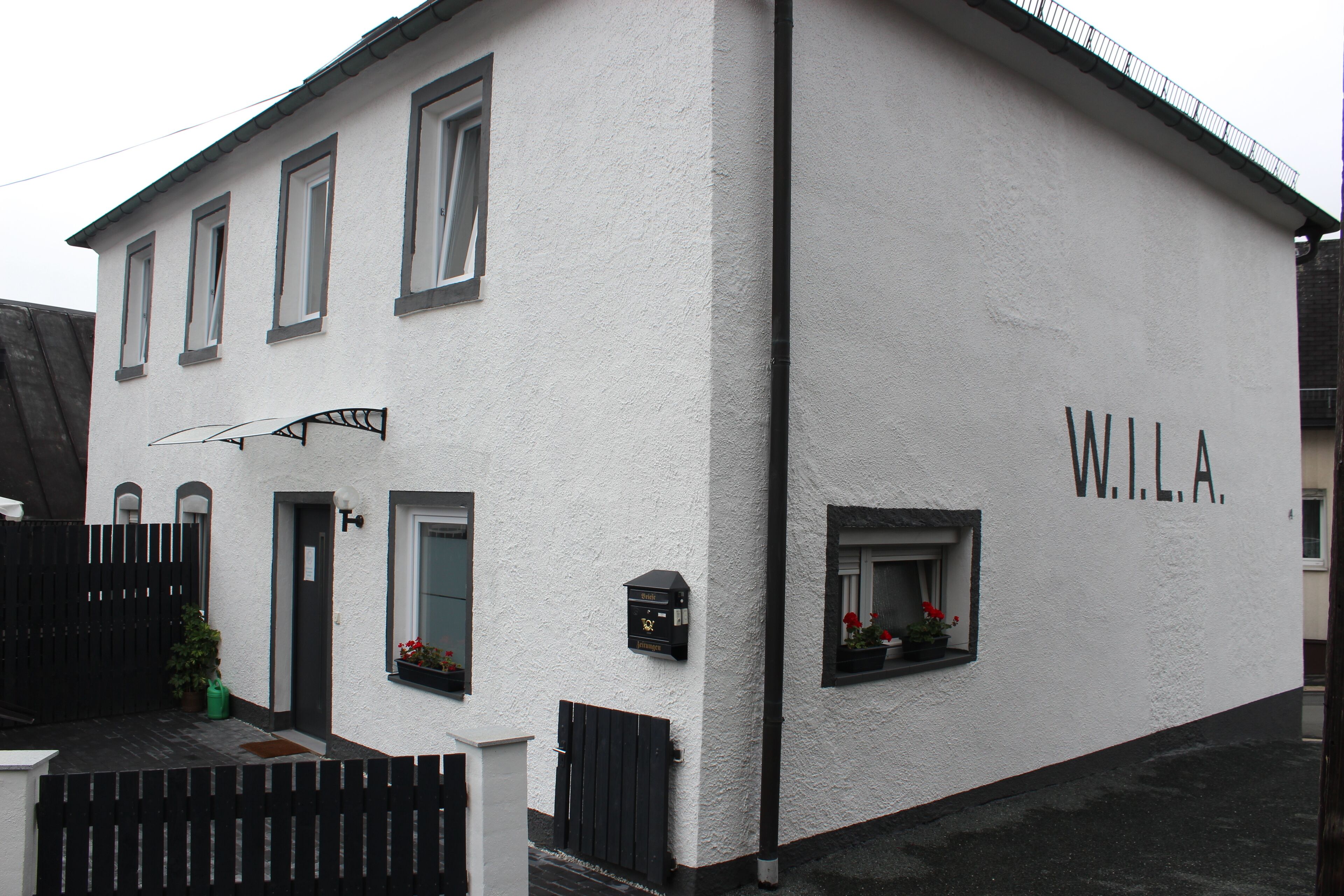 A white building with the name "WILA" on the side, a black gate, and a small window with flowers.
