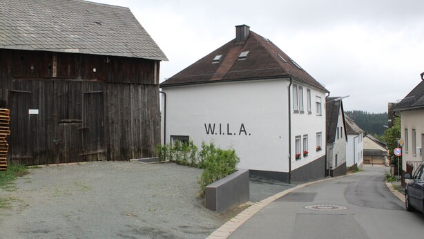 A street with a white building labeled "W.I.L.A." and a wooden barn. - W.I.L.A. 1 (Helmbrechts)