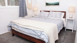 Een slaapkamer met een houten bed, een grijs-wit gestreepte dekbedovertrek, twee bedlampjes, een macramé wanddecoratie, een lijst met een landschapsafbeelding en een grijze gordijn.