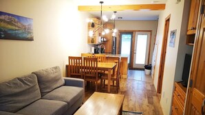 Un salon avec un canapé, une table basse en bois et des fauteuils. On y trouve également un coin repas avec une table et des chaises, et une cuisine à l’arrière-plan.