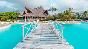 Una terraza de madera que conduce a una piscina turquesa, con construcciones de techo de paja al fondo.