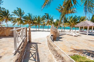 A beachside resort with a stone pathway leading to a seating area with lounge chairs and a thatched-roof structure.