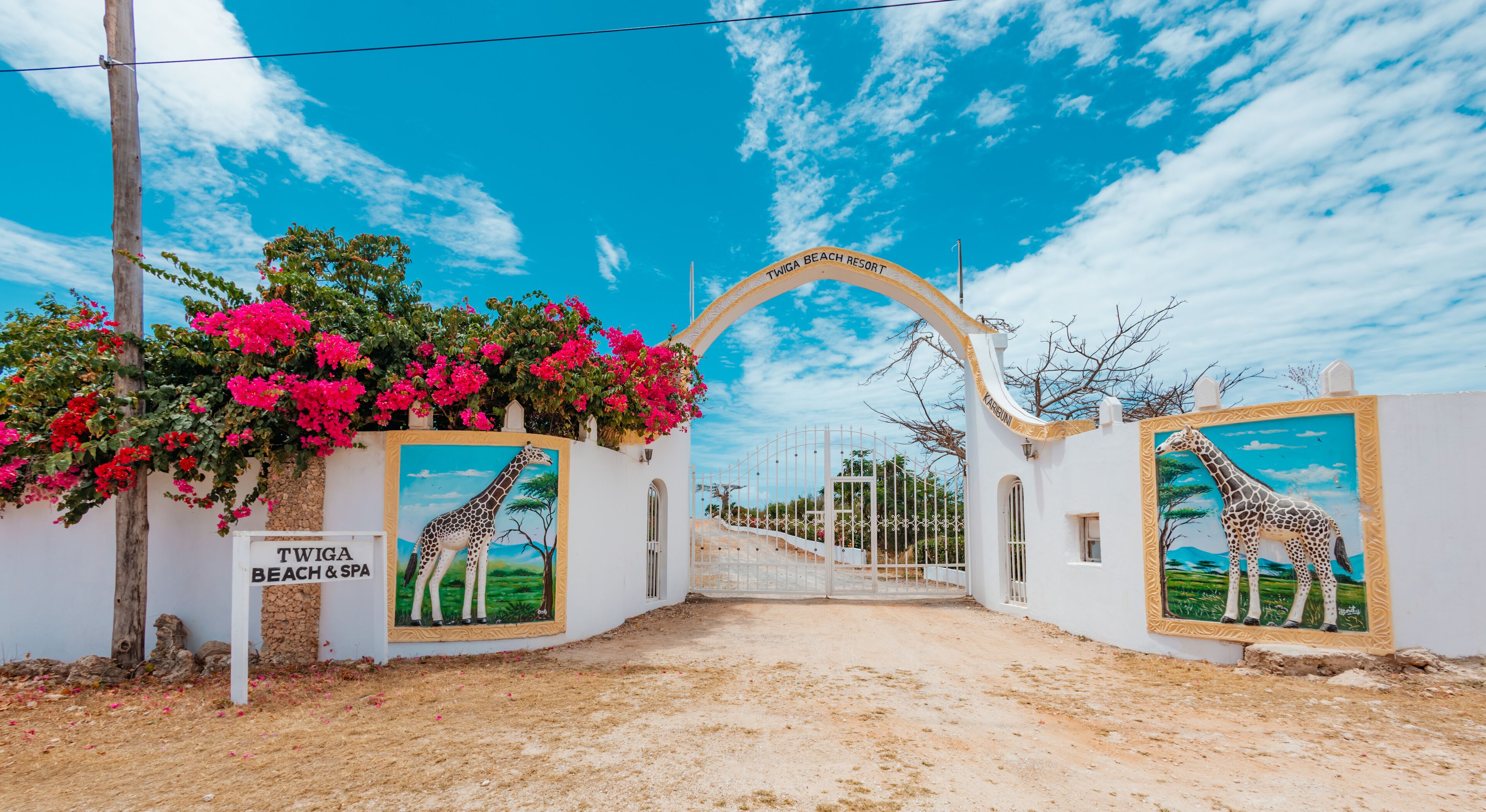 A white gate with a giraffe mural, a sign for Twiga Beach & Spa, and pink flowers.