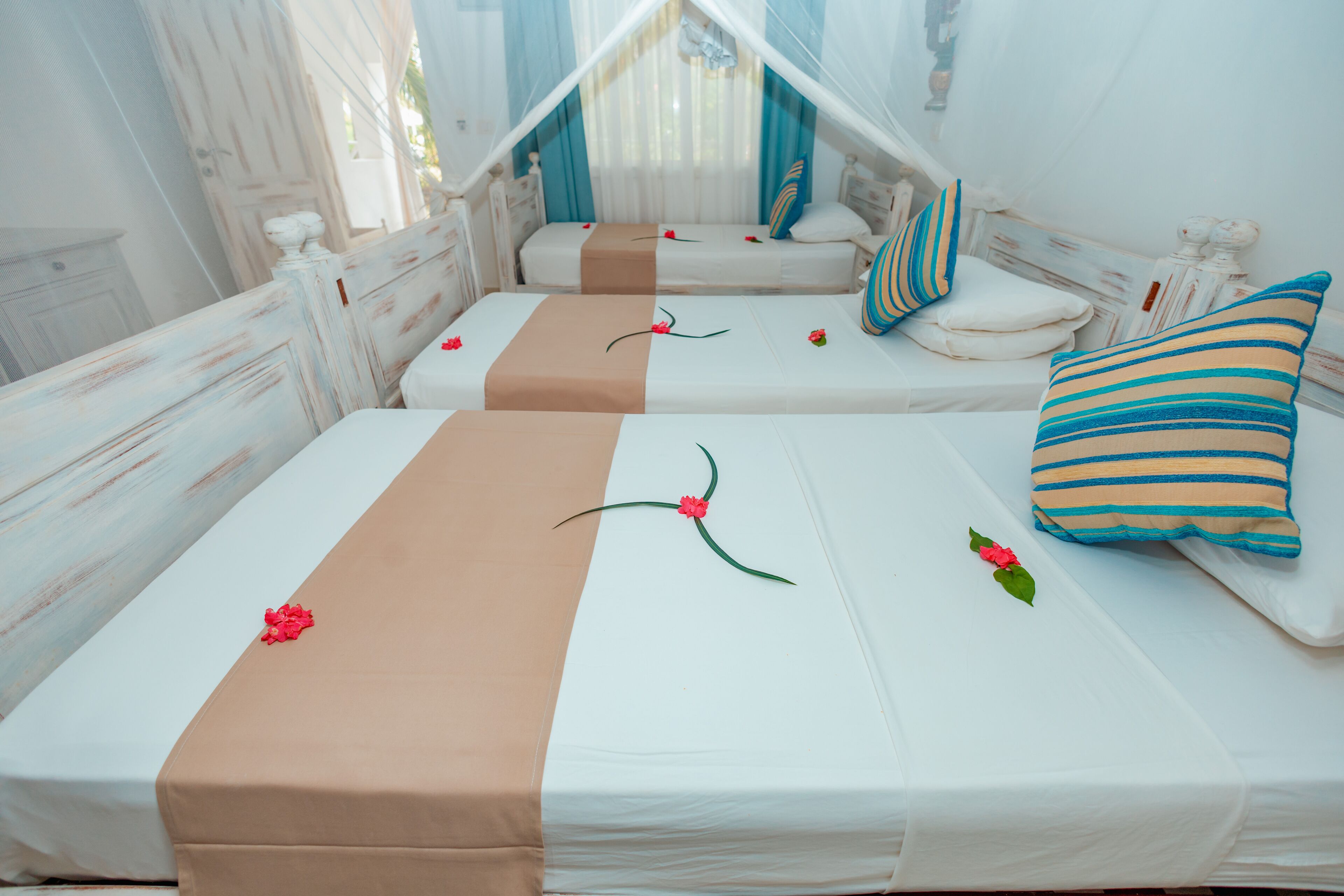 A room with two beds, each with a white mattress and a brown bed runner. There are decorative pillows and a canopy bed in the background.