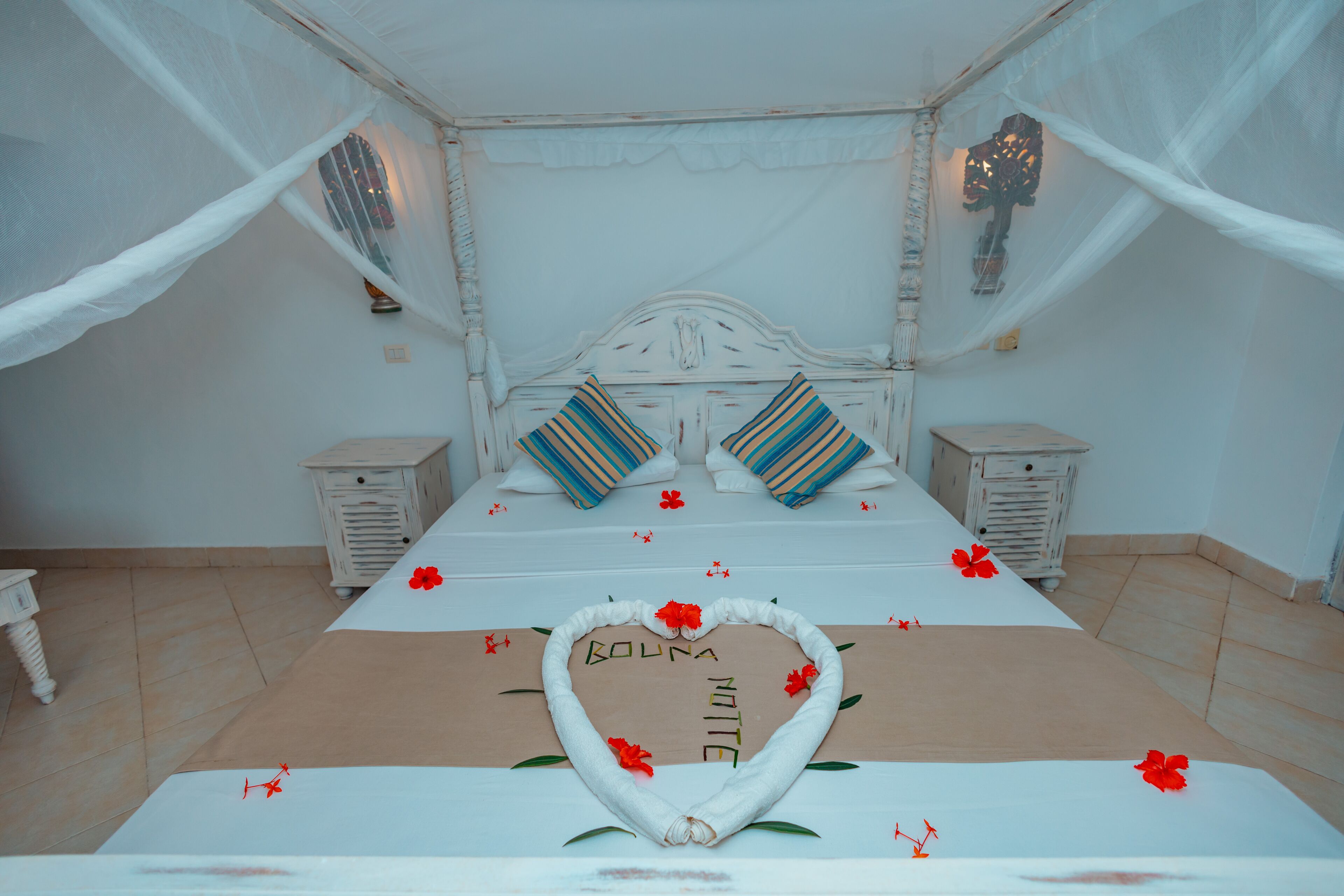 A bedroom with a canopy bed, two nightstands, a heart-shaped decoration on the bed, and a decorative wall piece.
