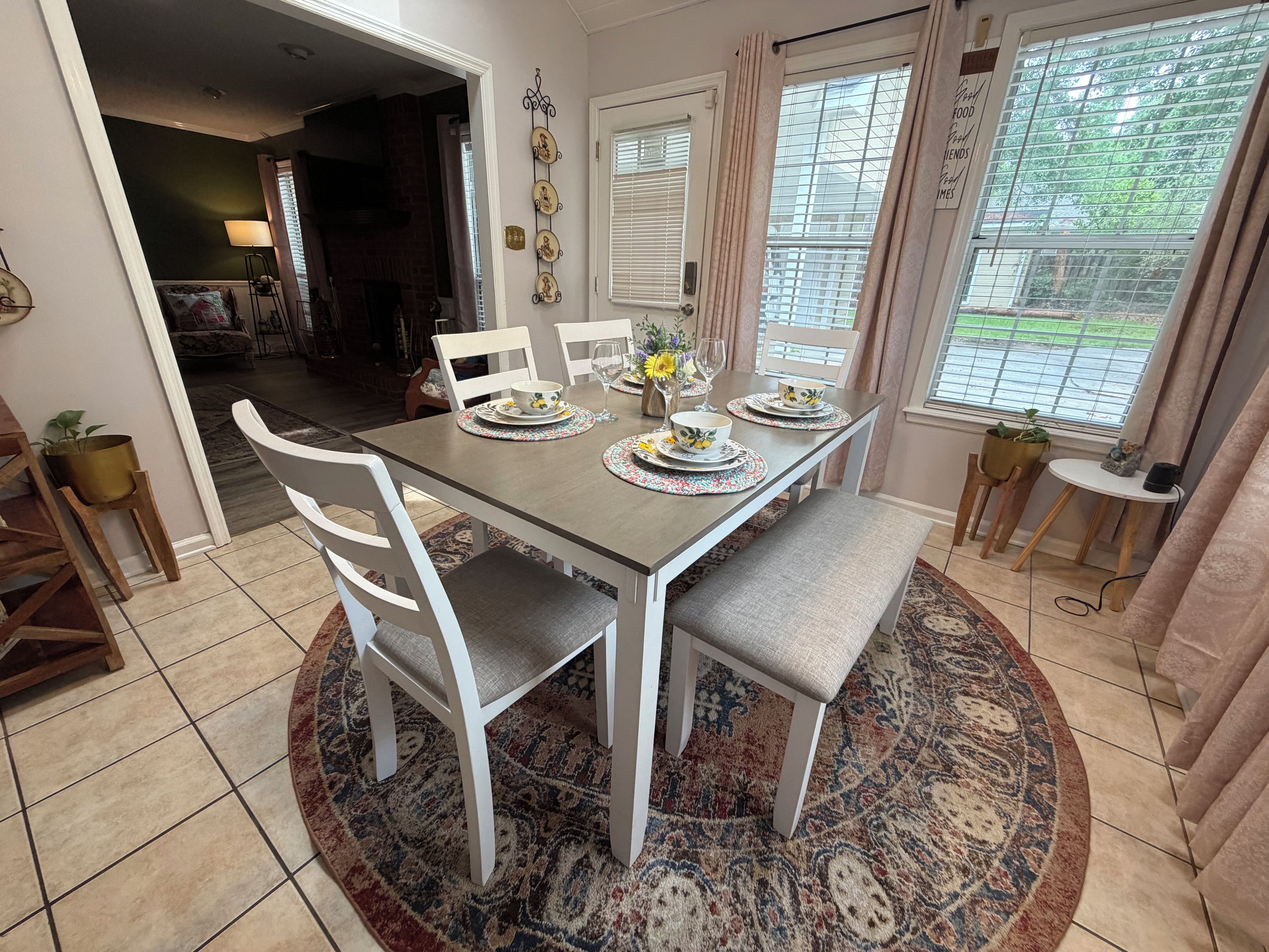A dining area with a table set for four, surrounded by white chairs, a patterned rug, and a view of the outdoors through a window with blinds.