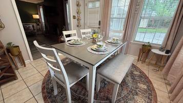 A dining area with a table set for four, surrounded by white chairs, a patterned rug, and a view of the outdoors through a window with blinds.