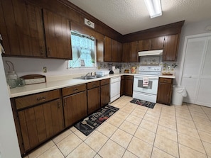 A kitchen with wooden cabinets, a white countertop, a window with a curtain, and a kitchen mat. - Spacious Peaceful Home 3BR 2BA,Quiet Neighborhood (Valdosta)
