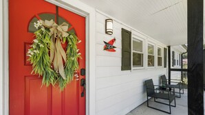Una puerta roja adornada con una corona, un pequeño espacio exterior con sillas y una mesa, y un cartel con el número de la casa.