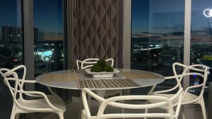 Un comedor moderno con una mesa redonda y cuatro sillas blancas, una pared decorativa y vistas a la ciudad a través de amplios ventanales.