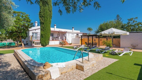Une piscine dotée d’une terrasse en pierre, entourée d’une pelouse verdoyante et agrémentée d’un parasol blanc.