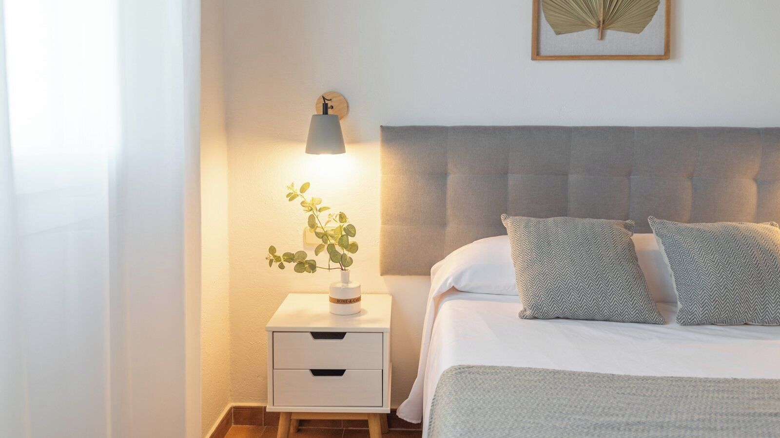 A neatly arranged bedroom with a bed, bedside table, lamp, and a framed plant on the wall.