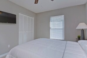 Un dormitorio con una cama, un televisor montado en la pared, una ventana con persianas y una lámpara de noche.
