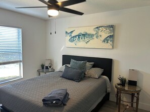 A bedroom with a bed, a ceiling fan, a window with blinds, a wall-mounted painting, a bedside table with a lamp, and a small round table with a plant.