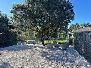An outdoor seating area with a gravel ground, surrounded by a fence, and a large tree providing shade.