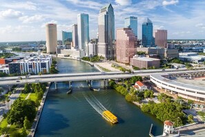 A cityscape with a river, modern buildings, and a yellow boat. - Casa Ahora! 3-Beds Walk to 7th Strip & TECO Trolly (Tampa)