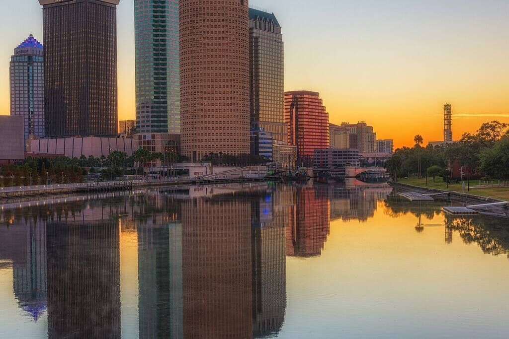 A city skyline with tall buildings reflected in a calm body of water during sunset.