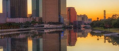 A city skyline with tall buildings reflected in a calm body of water during sunset.