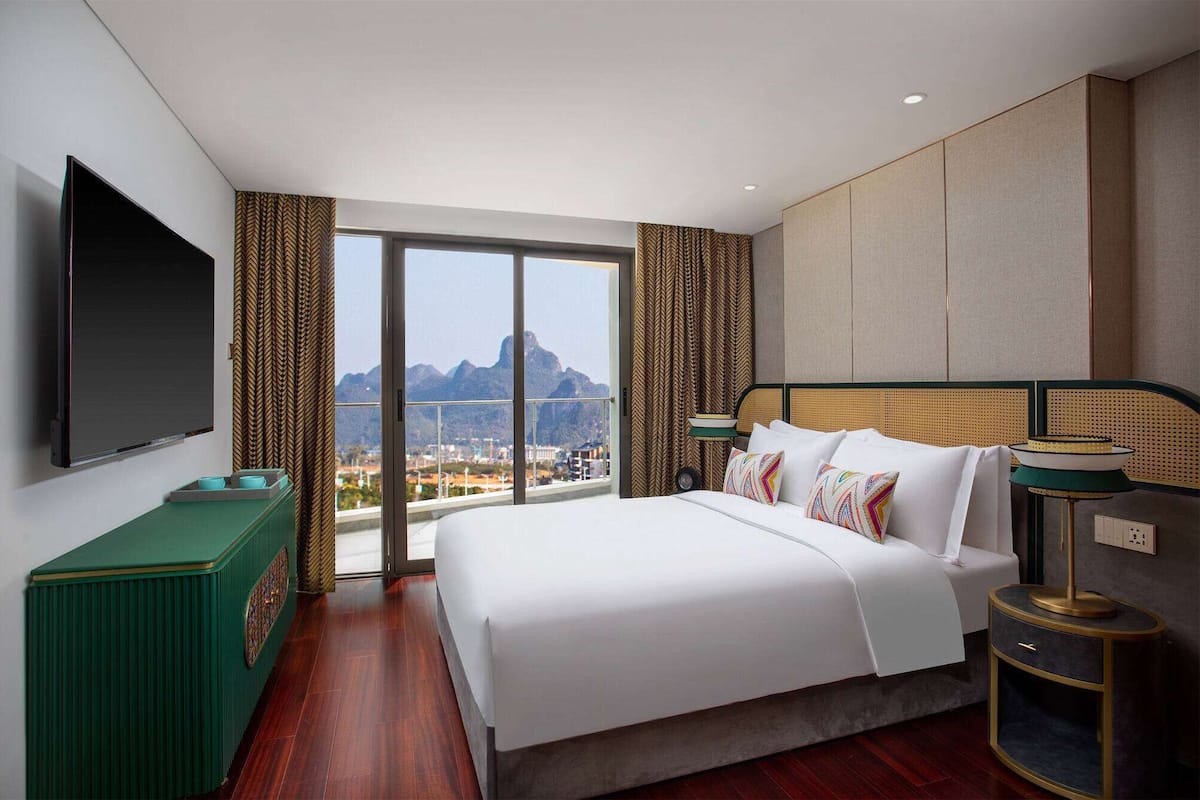 Habitación de hotel con una cama grande, un televisor, vistas a montañas y una mesita auxiliar verde.