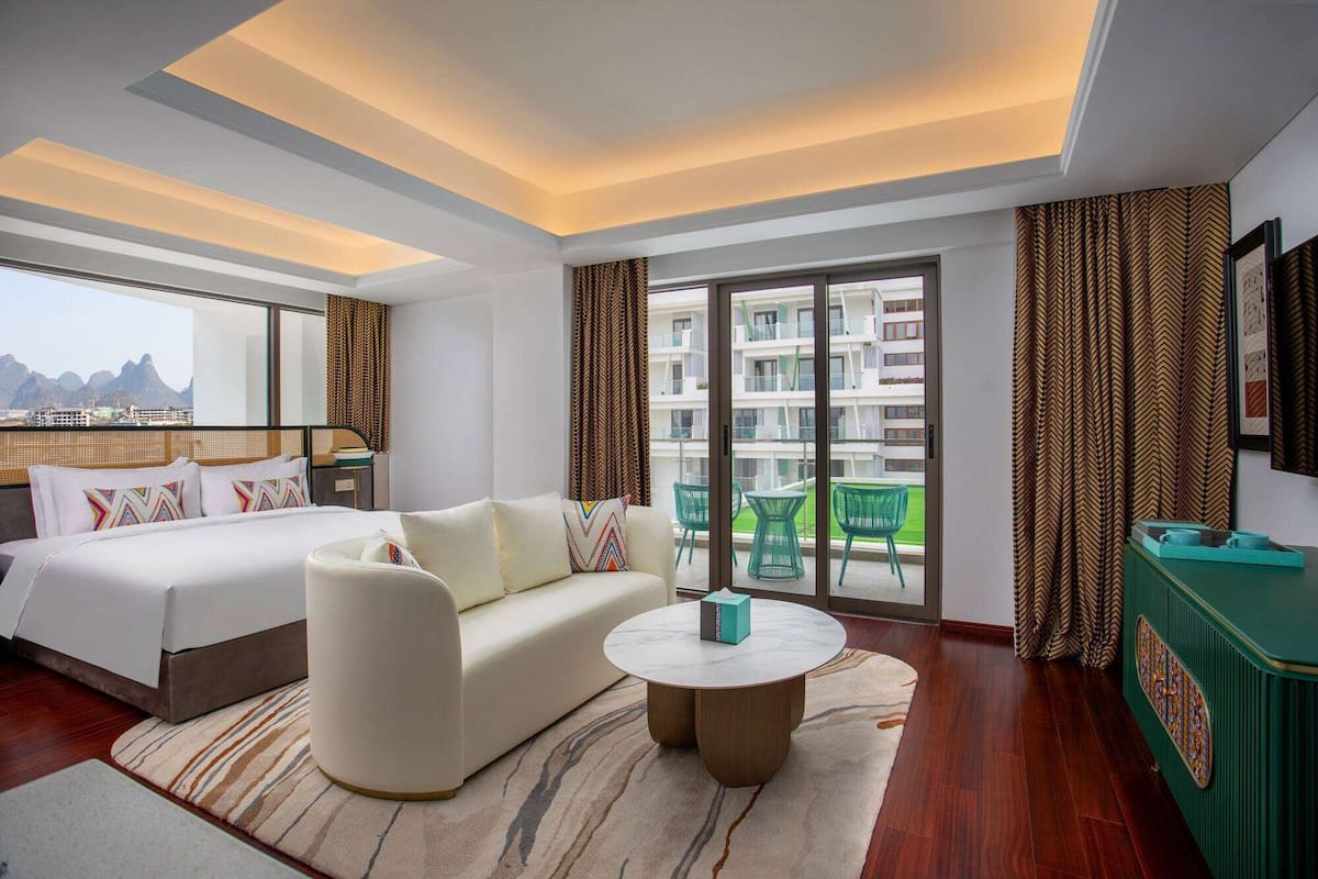 Una habitación de hotel moderna con una cama, un sofá, una mesita de centro redonda y un balcón con vistas a edificios y montañas.