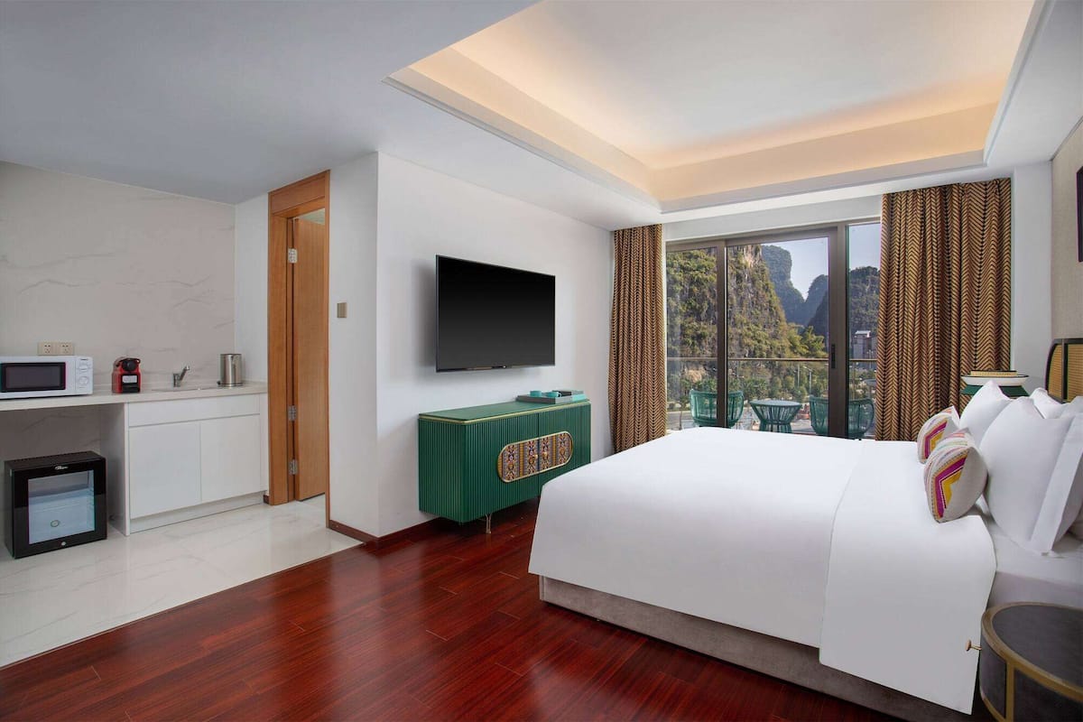 Habitación de hotel moderna con una cama grande, un televisor de pantalla plana, una kitchenette pequeña y un balcón con vista a la montaña.
