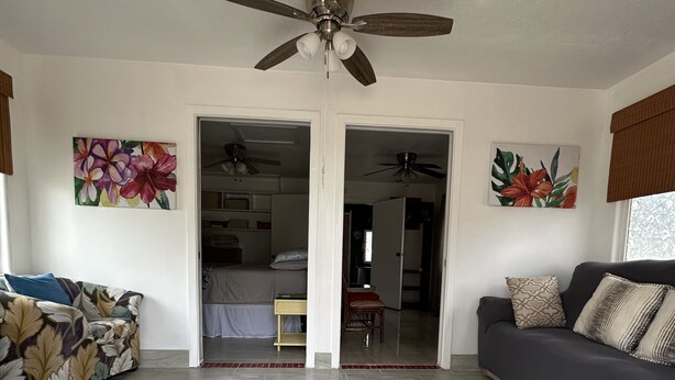 Un salón amplio con ventilador de techo, dos sofás, una mesa de centro y una mesa de comedor. Hay dos puertas que dan a otras habitaciones.