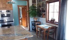 Une cuisine dotée d’un plan de travail en granit, de meubles en bois, d’un four encastré, d’une table à manger avec des chaises, d’une plante en pot et d’une fenêtre avec des rideaux.
