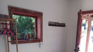 Une pièce avec un banc en bois, une fenêtre donnant sur un paysage verdoyant, une étagère ornée d’objets décoratifs et une porte menant à l’extérieur.