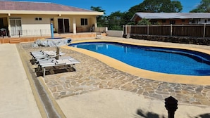 Una piscina con sillas de descanso, un patio de piedra y una casa al fondo.