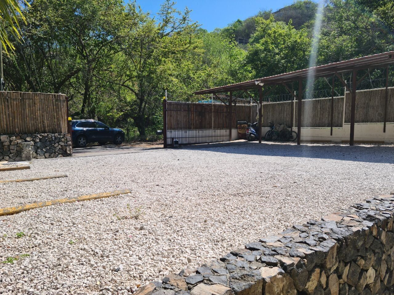 Un aparcamiento de grava con un muro de piedra, una valla de madera y una estructura cubierta.