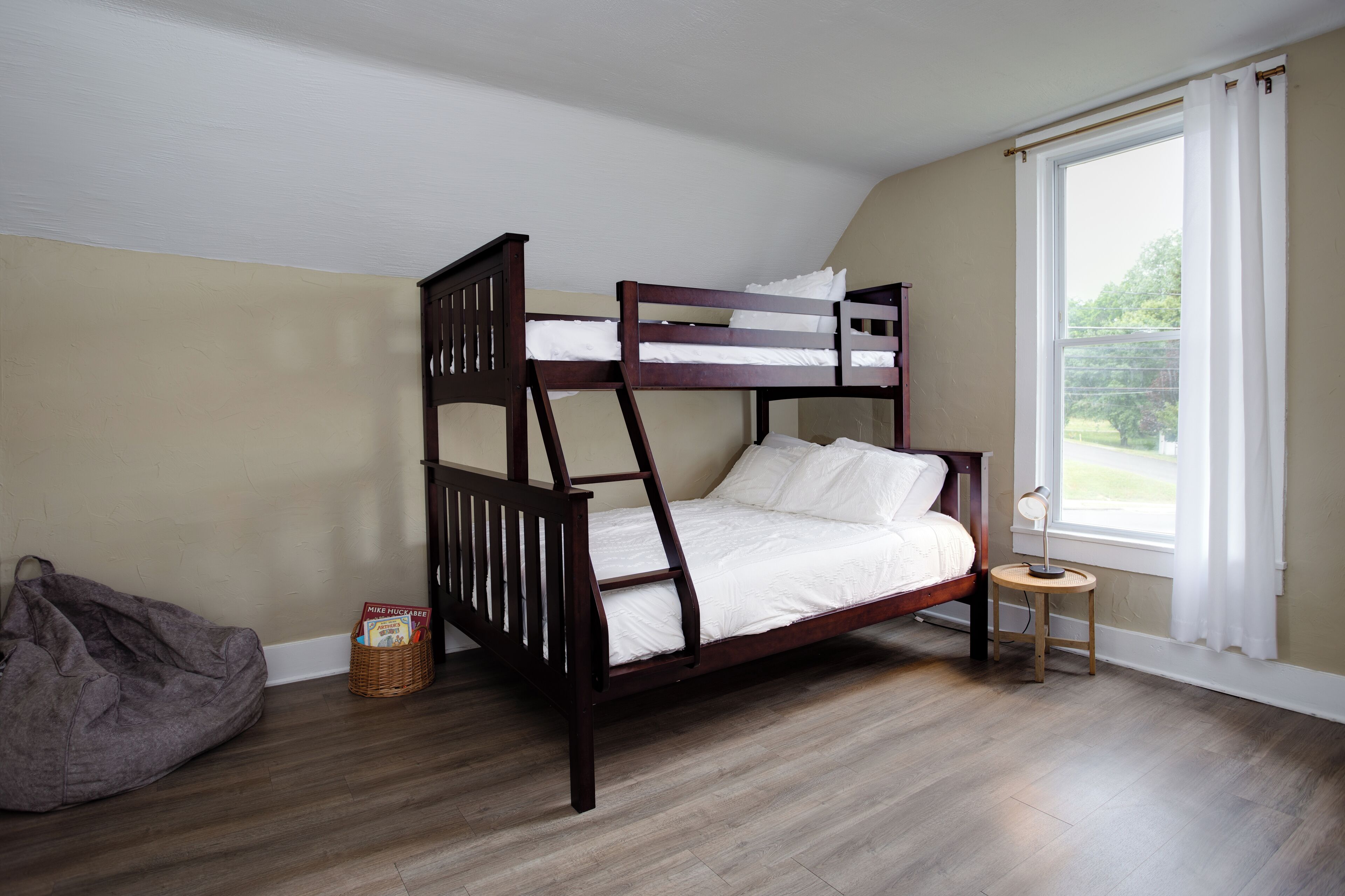 A bunk bed in a room with wooden flooring, a window with curtains, and a small table with a lamp.