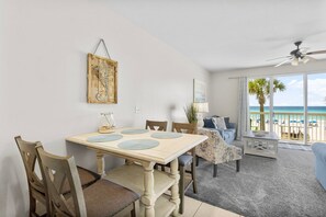 Un coin repas avec une table et des chaises en bois, un ventilateur de plafond et une vue sur la plage grâce à de grandes fenêtres.