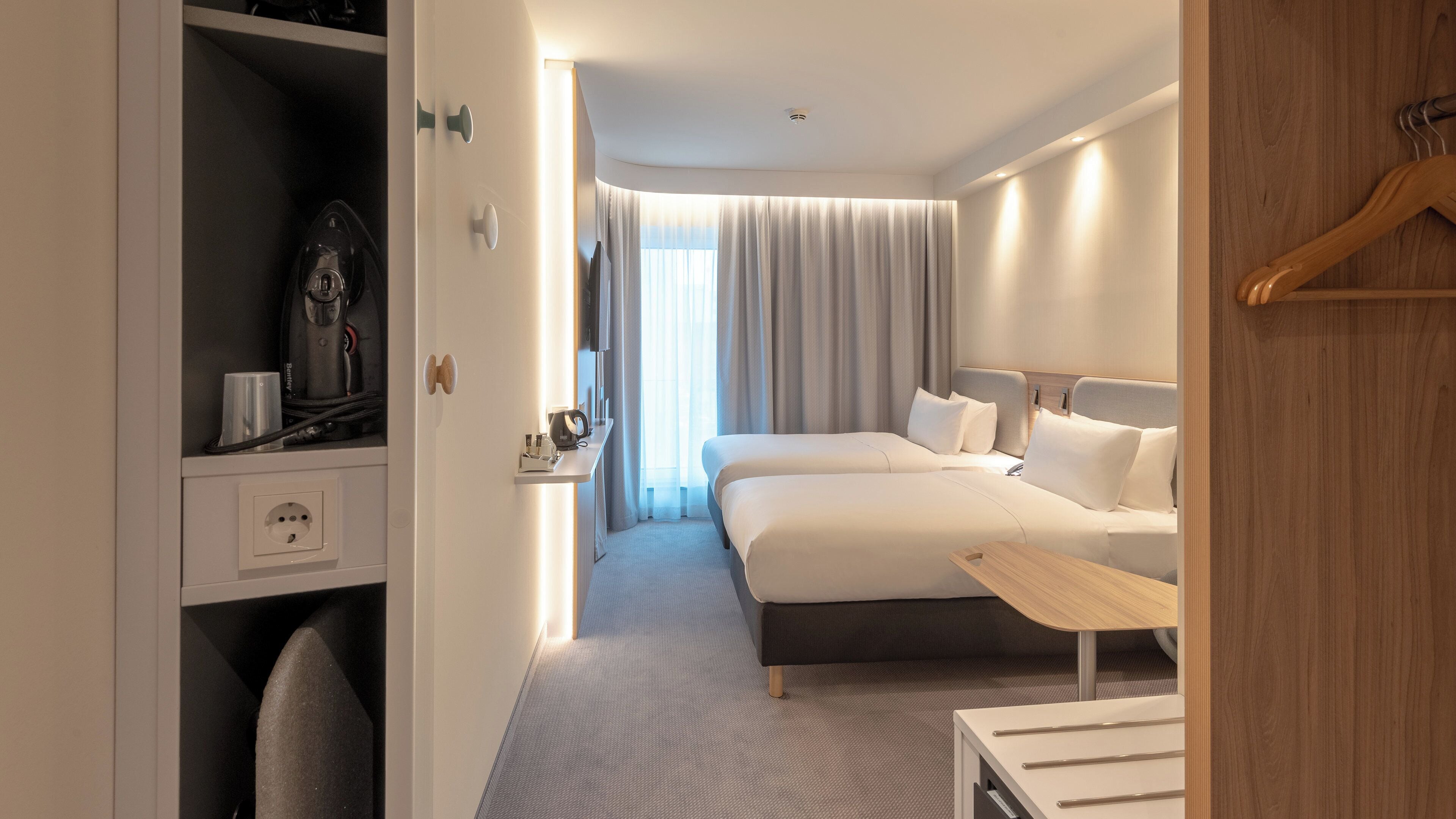 A hotel room with a bed, bedside tables, a desk with a chair, a wardrobe, and a view of the outside.