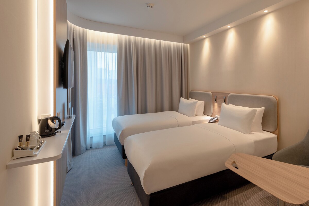 A hotel room with two beds, a desk, a chair, a TV, and a window with curtains.