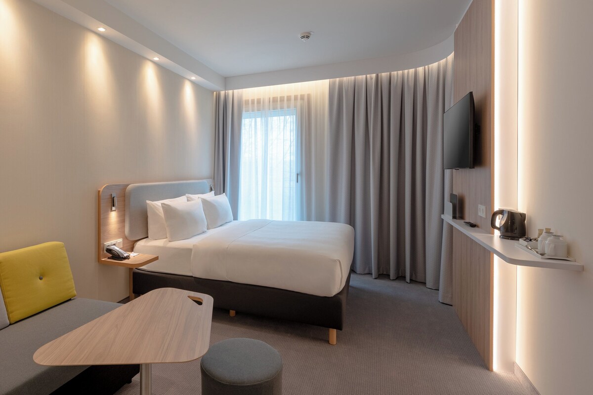 A modern hotel room with a bed, a sofa, a small table, and a TV.