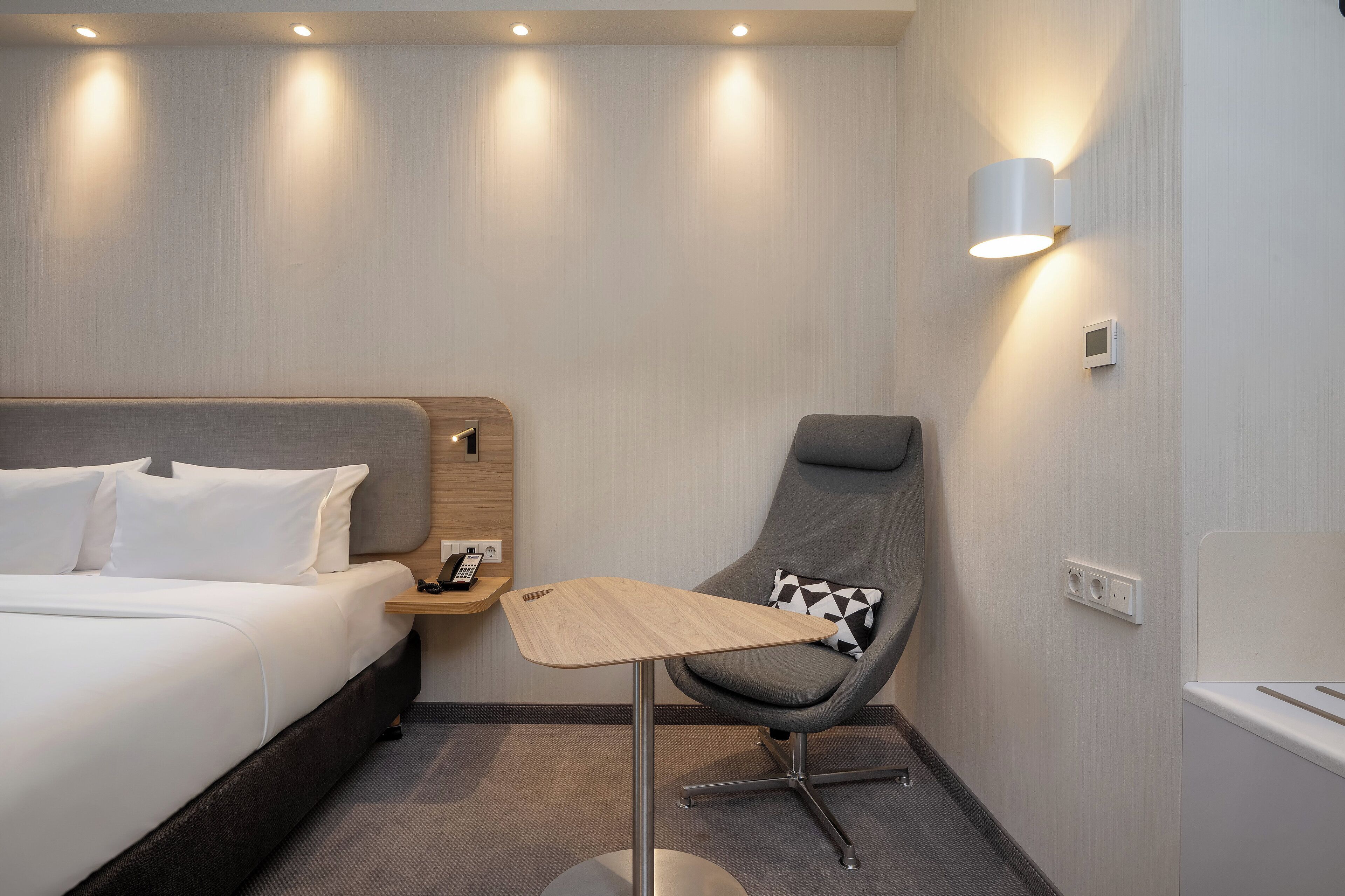 A hotel room with a bed, a desk, a chair, and a wall-mounted light fixture.