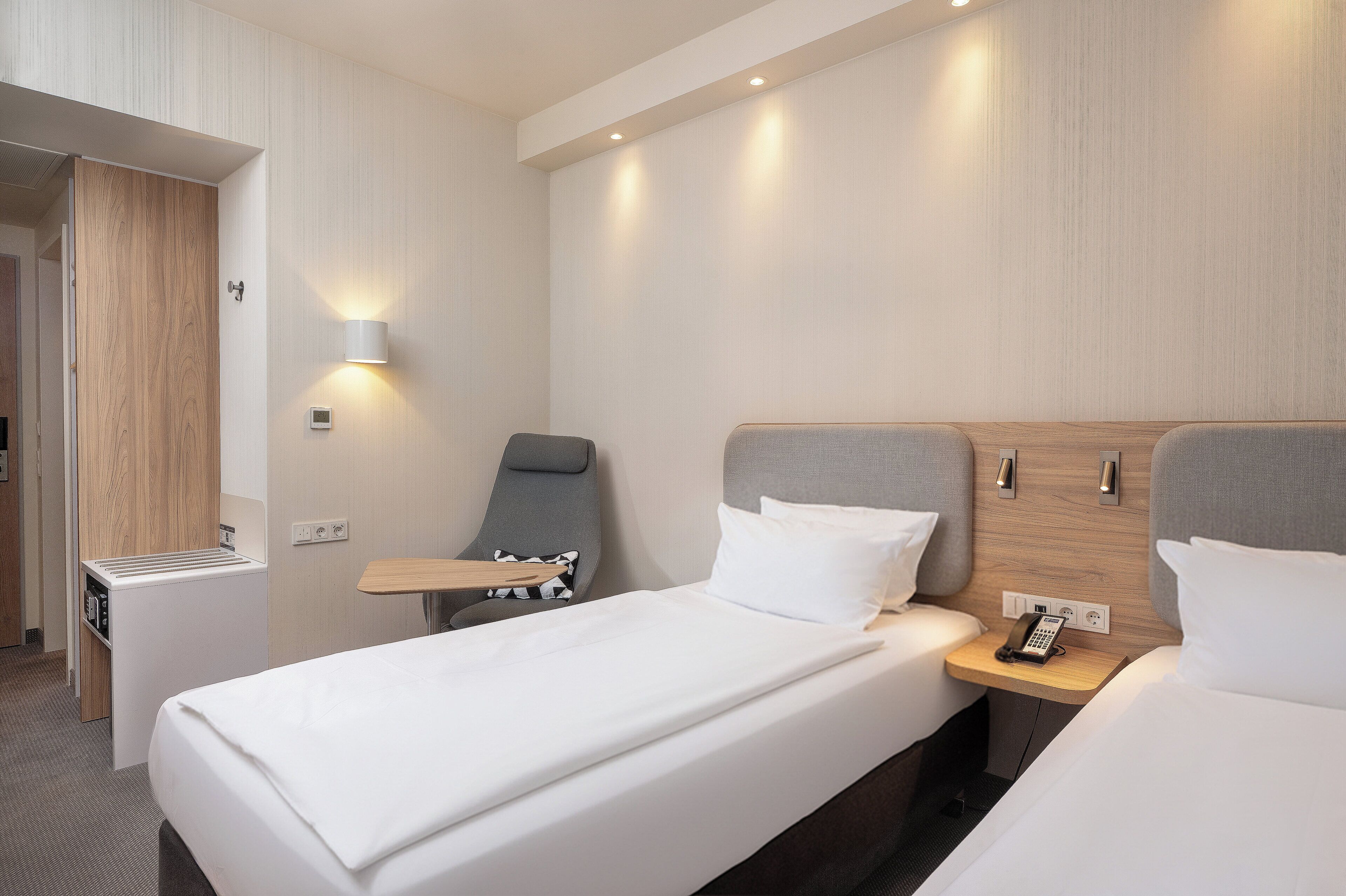 A hotel room with a bed, bedside table, chair, and a small kitchenette.
