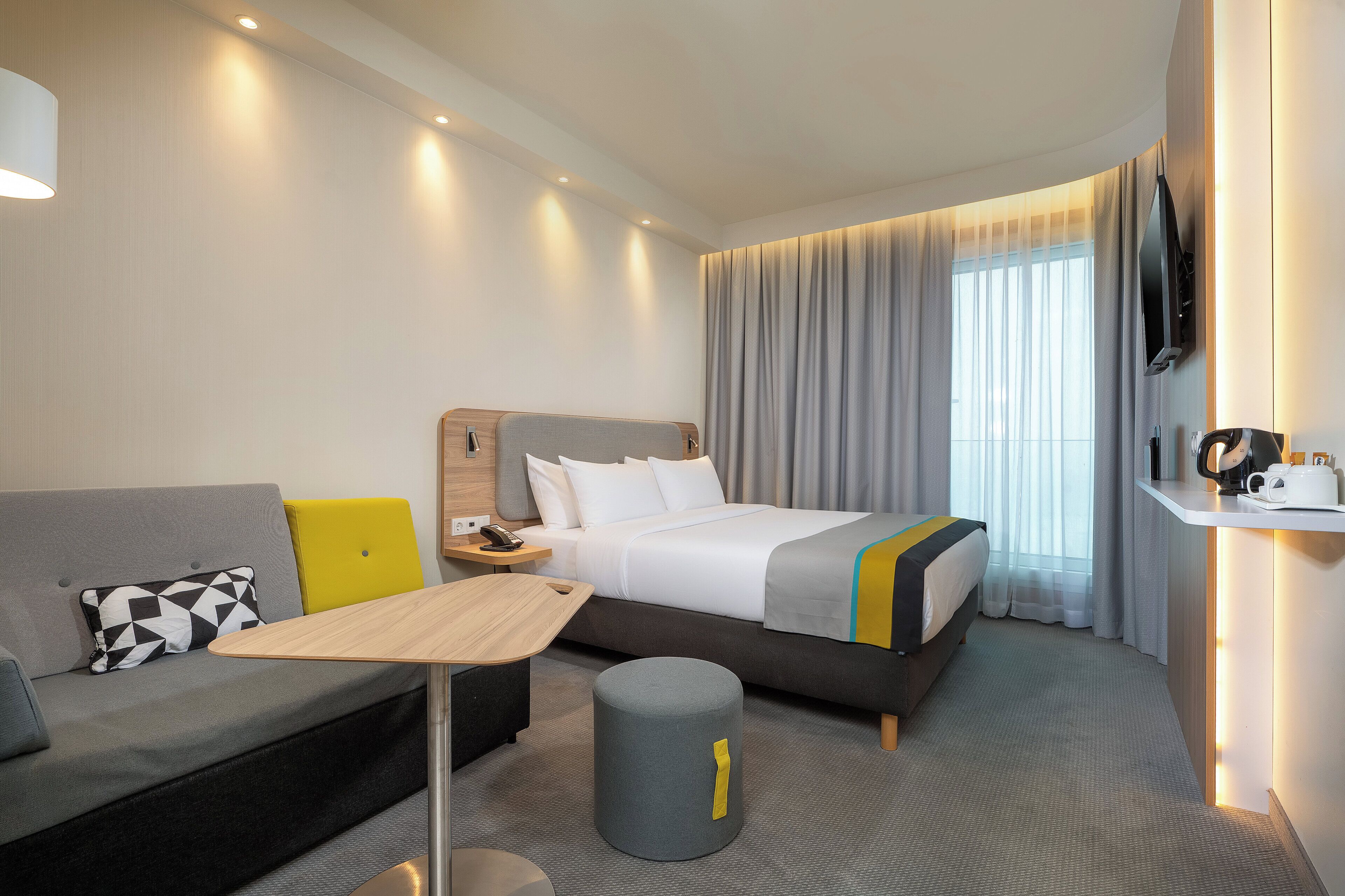 Une chambre d’hôtel moderne équipée d’un lit, d’un canapé, d’une table de chevet, d’une lampe et d’un petit tabouret.