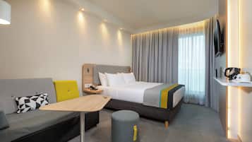 A modern hotel room with a bed, sofa, bedside table, lamp, and a small stool.