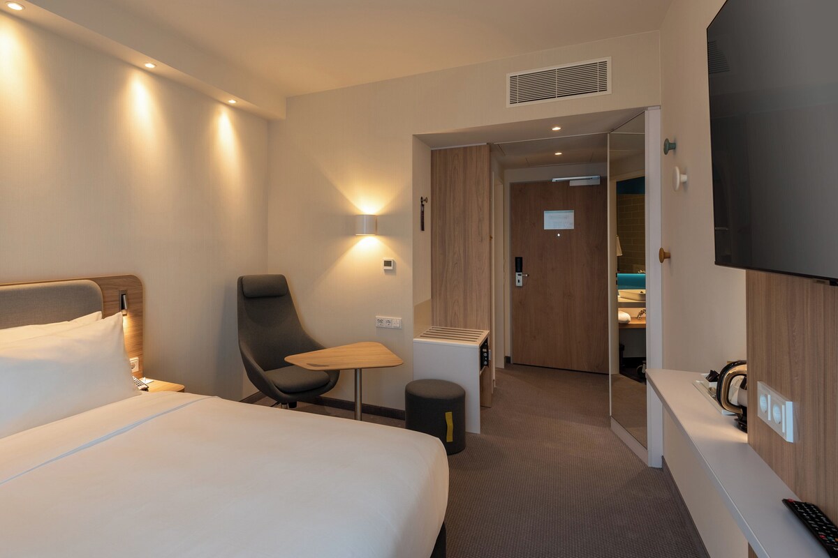 A hotel room with a bed, a chair, a small table, a TV, and a bathroom visible through an open door.