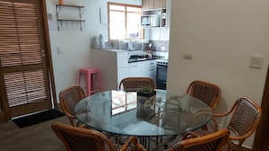 A dining area with a glass table surrounded by wicker chairs, a pink stool, and a kitchen in the background. - Cozy house with 2 garages and 2 bedrooms. (SP)