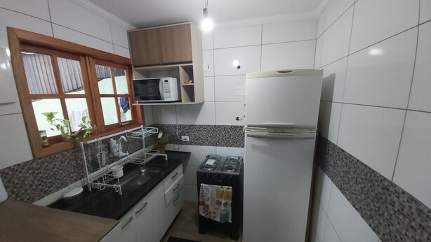 A compact kitchen with white cabinets, a microwave, a refrigerator, a gas stove, and a window with curtains. - Cozy house with 2 garages and 2 bedrooms. (SP)