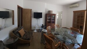 A living room with a glass-top dining table surrounded by wicker chairs, a sofa with a patterned cushion, a television mounted on the wall, and a cabinet with glass doors. - Cozy house with 2 garages and 2 bedrooms. (SP)