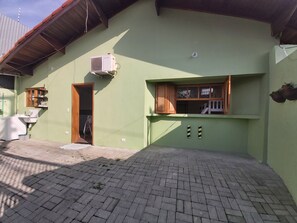 A green building with a tiled patio, a window with a wooden frame, and a small shelf with items. - Cozy house with 2 garages and 2 bedrooms. (SP)
