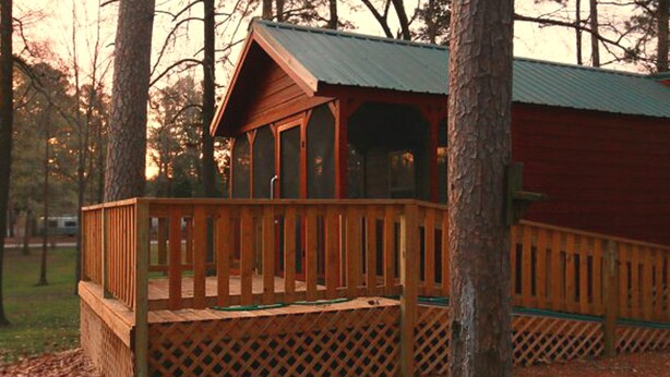 A wooden deck with a lattice railing, a covered porch, and a red building in a wooded area. - Birdsong Resort Marina Lakeside RV (Camden)